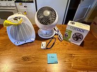 All three appliances side by side on table: heater fan left, Vornado orb fan center with remote and lanyard, O2Cool boxed fan right