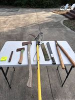 All seven tools laid out on table: wedge, hatchet, axe, pole pruner, machete with sheath, two wooden clubs