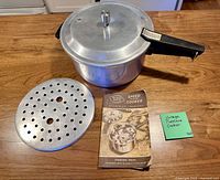 Mirro-Matic aluminum pressure cooker with lid, steamer insert and manual displayed on countertop