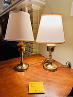 Pair of brass candlestick table lamps with white fabric shades shown on wooden surface