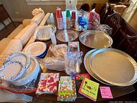 Tabletop view showing all disposable party supplies including plastic cups, paper plates, plastic cutlery, napkins, and aluminum serving trays