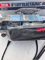Close view of Ryobi BE-321 sander with dust bag and model label, box partially visible