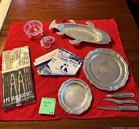 All items in lot arranged on red cloth: boxed seafood set, two pewter plates, fish platter, two knives, glass candy dish with lid, two Camden Maine tea towels