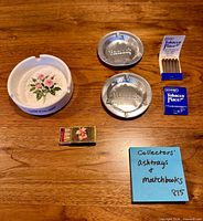 Overview of one ceramic ashtray, two metal ashtrays, two matchbooks, one matchbox on wood surface