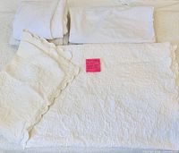 Folded white embroidered bedspread, scalloped pillow shams, fitted sheets and other bedding pieces displayed