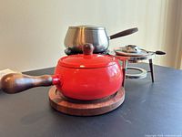 Group view of red enameled pot on wood trivet, stainless pot behind, burner stand with insert at right