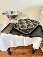 Stacked muffin pans, baking sheets and stainless steel colander displayed on table