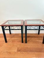 Both square end tables side by side showing glass tops, wood trim, black metal frames with scroll accents, and floor context