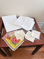 Folded white tablecloth, stack white napkins, assorted printed and embroidered handkerchiefs on table