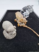 Tray showing three pig figurines: grey resin, tan pull-toy on wheels, clear glass
