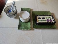 All items laid out: stack of four bowls with two spoons, three white dishes, two green plates, black fruit plate