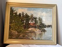 Front view of framed oil painting depicting lakeside cottage and trees