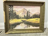 Front view of oil painting showing mountain scene, trees, meadow, reflective water; ornate gilt frame with left side damage visible