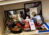 Shelf view of assorted Asian decorative items including porcelain figurine, lacquer boxes, framed prints, cloisonné pieces