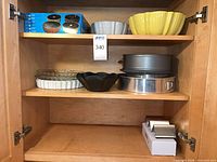 Cabinet shelf showing boxed mini Bundt pan, two large ceramic bowls, pie plates, springform pans and Bundt pan