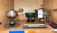Cabinet view showing two stainless pots, green ceramic pot, stands, burners, and fork trays