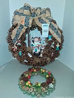 Large pine-cone wreath with burlap ribbon and clay figurines hanging above small fruit wreath