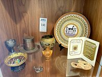 Group shot of coin certificate, Rhodes plate, urn, painted glass dish, metal goblet, inlaid container, trinket, terracotta fragment