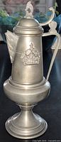 Full left profile of lidded pewter tankard showing crown motif, finial, handle, pedestal base