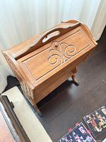 Overall view of wooden sewing cabinet showing folding lid, carved handle and scroll motif, on caster feet