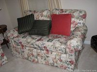 Floral print loveseat with three pillows: two dark green striped and one solid red. Loveseat upholstered in floral fabric with shades of pink, red, green, and cream. Plush cushions with skirted bottom and rounded arms.