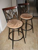 Two bar stools featuring metal frames with a curved backrest pattern and wood trim at top backs with tan velour seats on a tiled floor.