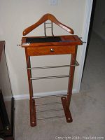 Full view of single wooden valet stand with metal rods for hanging and small drawer, placed against wall on carpeted floor.