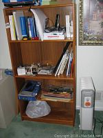 Wood bookcase filled with books, office supplies, and miscellaneous items; adjacent Lakewood model 7096 space heater visible