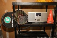 Photo showing two tennis rackets, a handball racket, Brookstone CD player, and orange cones on a shelving unit.