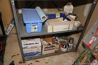 Shelf with multiple boxes of Rapid Set Cement All, a blue plastic toolbox, some paint cans, and assorted tile installation supplies