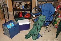 Three folding chairs with blue fabric and metal frames, along with carrying cases, purple and white cooler, and blue folding crate placed on the floor in a garage setting.