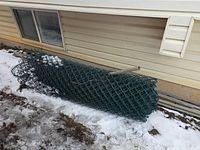 Rolled green chain link mesh with a rail inside, lying beside house foundation on snow
