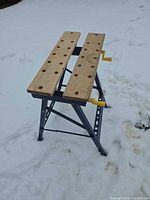 Three-quarter view of blue folding sawhorse workbench in snow showing wooden jaws and crank handles