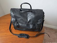 front view of black leather carry-on with flap, buckles, handles, strap