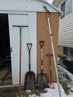All five garden tools leaning against shed: rake, two shovels, edger, pole pruner