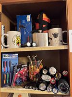 Entire cabinet shelf view showing flashlights, mugs, hip flask and insulated bottle
