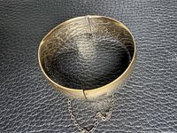 Top view of oval hinged gold-tone bangle bracelet showing clasp and safety chain