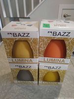 Front view of four boxed Bazz Lumina lamp shades showing colors yellow, orange, grey
