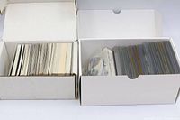 Two open white card storage boxes, left containing loose cards, right containing sleeved cards