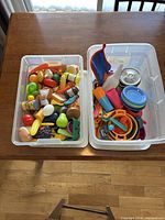 Two clear bins; left bin filled with assorted plastic play food, right bin with toy dishes and utensils