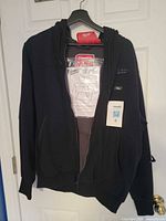 Front view of black Milwaukee M12 heated hoodie on hanger with tags and manual visible