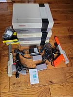 Three NES consoles stacked with zappers, RF switch boxes, AC adapters and cables visible