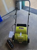 Front view of Sun Joe scarifier/dethatcher with handle, wheels, bag, power cord and accessories on floor