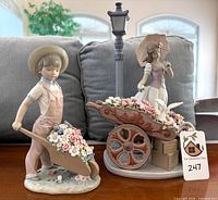 Two Lladró figurines: boy with wheelbarrow of flowers and woman with parasol behind flower cart
