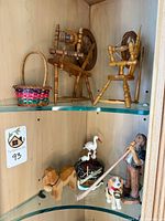 Assortment of wooden spinning wheels, woven basket, dog figurines, stork figurine, and alpine man figure displayed on glass shelves