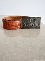 Coiled brown leather belt beside brass Colt Revolvers buckle front view