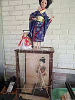 Three dolls shown: large blue kimono, small red kimono, medium doll in glass case; view of damaged stand