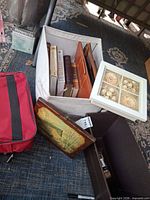 Lot overview showing books in fabric bin, red carry bag, framed decor, wood plaque, brown box
