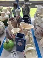 Assorted vases displayed including blue jasperware-style, green round, cream basket-form, ribbed with appliqué, cobalt blue, cream scalloped, white hobnail, floral appliqué, green textured, white grape motif and ceramic bell