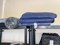 Two folded blue quilted moving blankets and one rolled grey pad on shelf